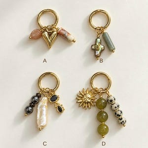May include: Four gold-toned metal rings labeled A, B, C, and D, each featuring various charms. Ring A has a heart, a peach bead, and a striped cylinder. Ring B has a cross and a green cylinder. Ring C has dark beads, a pearl, and black stones. Ring D has a sun, green beads, and spotted beads.