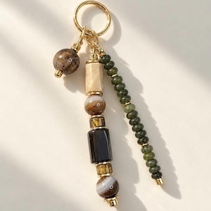 May include: A gold-toned metal keychain featuring a central vertical strand of assorted beads in shades of tan, brown, and black, flanked by a single round brown bead on one side and a long vertical strand of small, round, dark green beads on the other.