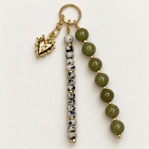 May include: A gold-toned metal ring charm featuring three dangling elements: a small textured heart, a strand of speckled dalmatian jasper beads with gold spacers, and a strand of smooth olive green round beads separated by small gold beads.