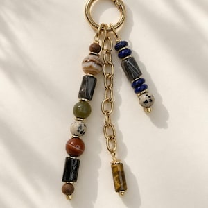 May include: A gold-toned metal keychain featuring a circular ring attached to three dangling strands. Two strands consist of assorted multi-colored stone beads in spherical and cylindrical shapes, while the third is a gold-toned metal chain.