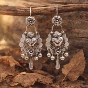 May include: A pair of silver-toned dangle earrings. Each earring features a floral top, a heart-shaped centerpiece, and dangling teardrop and ball accents. The earrings are displayed against a natural, blurred background.