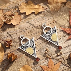 Puede incluir: Pendientes colgantes plateados con un diseño geométrico. Cada pendiente presenta una forma triangular con un patrón texturizado, un círculo dorado y una pequeña gema roja. Los pendientes se exhiben sobre una superficie de madera con hojas de otoño.