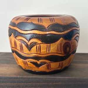 May include: A small, rounded wooden bowl featuring a polished finish with alternating bands of natural light brown wood and dark brown stained sections. The surface is decorated with carved horizontal wavy lines and vertical hatch marks.