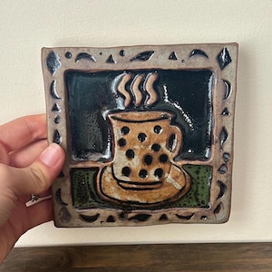 May include: A square ceramic tile featuring a raised relief of a polka-dot mug on a saucer with steam rising. The background is dark green, and the border is decorated with small black geometric shapes, including diamonds and crescent moon symbols.