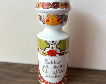 May include: A white ceramic canister with a lid featuring a floral pattern and a stylized face. The body displays green and red floral motifs and the text "Sukker på kake vil alltid smake" in black script. The design includes a decorative border.