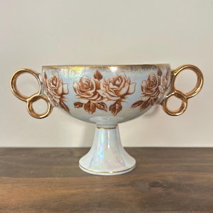May include: A footed porcelain bowl with an iridescent finish, featuring two gold-toned handles and a repeating pattern of brown roses around the exterior. The rim and base are accented with gold trim, and the bowl has a wide, shallow basin.