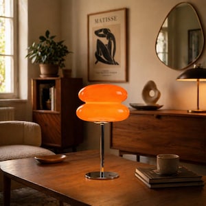 Puede incluir: Lámpara de mesa naranja con forma de doble burbuja y base cromada. La lámpara está sobre una mesa de madera en una habitación con un estampado de Matisse y un mueble de madera. Diseño de mediados de siglo.