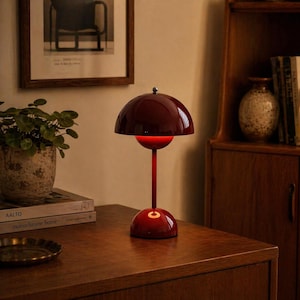 Può includere: Una lampada da tavolo color bordeaux con un paralume a forma di cupola e un gambo sottile. La lampada ha una base circolare e una piccola luce rossa brillante. Si trova su una superficie di legno, accanto a una pianta in vaso e a un quadro.