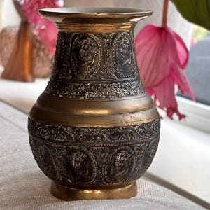 Op de afbeelding: Decoratieve messing vaas met een gedetailleerd, reliëf ontwerp. De vaas heeft een donkere, verouderde afwerking met gouden accenten aan de bovenkant, basis en midden. Het reliëfpatroon bevat figuren en bloemmotieven.