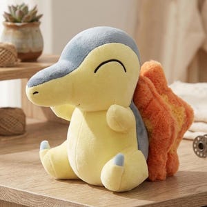 Puede incluir: Un peluche de Cyndaquil en posición sentada. El juguete presenta un cuerpo amarillo, detalles azules y grises, y una llama naranja en la espalda. Tiene una cara sonriente y pequeños pies azules.