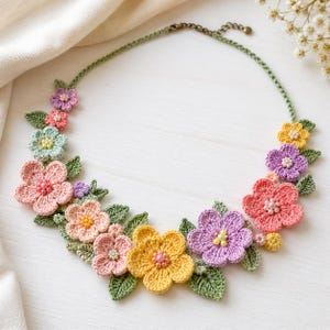 Può includere: Una collana all'uncinetto fatta a mano con fiori colorati nelle tonalità del rosa, giallo, viola e verde. Il design floreale è accentuato da foglie verdi e una delicata catena.