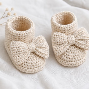 May include: A pair of cream-colored, crocheted baby booties. Each bootie features a large bow on the front. The booties are made of a soft, textured yarn and are displayed on a white, textured surface. A sprig of dried flowers is in the background.