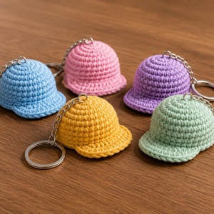 Puede incluir: Cinco llaveros de gorra de béisbol tejidos a ganchillo en azul claro, rosa, amarillo, morado y verde claro. Cada gorra presenta una corona redondeada y una visera corta, unida a una anilla de metal plateado mediante una cadena corta.