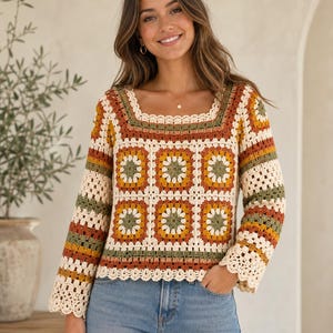 Puede incluir: Un suéter de crochet con cuello cuadrado y diseño patchwork. El suéter presenta una base color crema con cuadrados en verde oliva, amarillo mostaza y acentos color óxido. Las mangas tienen un borde acampanado y festoneado. La modelo lleva vaqueros azules.