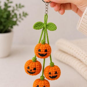 Può includere: Un portachiavi fatto a mano a tema Halloween con quattro zucche arancioni all'uncinetto con facce sorridenti nere. Le zucche sono sospese a steli e foglie verdi, attaccate a un portachiavi argentato. Un accessorio festivo.