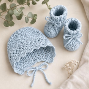 Puede incluir: Conjunto de gorro y patucos de bebé de ganchillo azul claro. El gorro tiene un borde festoneado y lazos bajo la barbilla. Los patucos tienen un detalle de lazo en el tobillo. El conjunto está hecho de hilo suave.