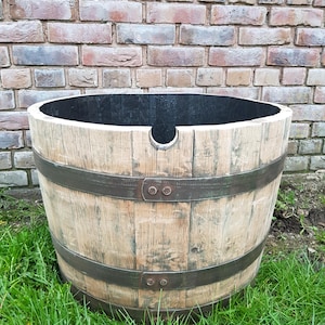 May include: A large, round wooden barrel planter featuring light-coloured vertical staves and two dark, weathered metal bands secured with rivets. The interior of the barrel is painted black, and a small semi-circular notch is cut into the top rim.
