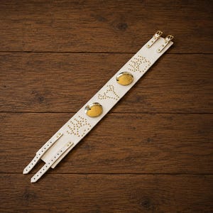 May include: A long, white leather cuff bracelet featuring two gold-toned circular metal accents and decorative gold-toned stud patterns. The cuff includes two adjustable straps with gold-toned buckles and multiple punched holes for secure fastening.