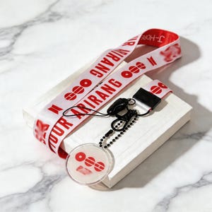 Könnte beinhalten: Ein weißes Schlüsselband mit rotem Text und Grafiken, darunter die Wörter "YOUR ARIRANG" und "J-HOPE". Das Schlüsselband hat einen schwarzen Verschluss und einen transparenten, kreisförmigen Anhänger mit passenden roten Grafiken. Das Schlüsselband liegt auf einer weißen Box.