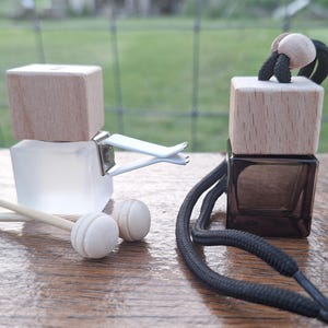 May include: Two car air fresheners with wooden tops and glass containers. One is clear with a clip, the other is dark with a black cord. Two small wooden sticks and two round wooden balls are in front.