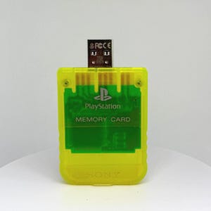 May include: A yellow PlayStation memory card with a USB adapter. The card has the PlayStation logo and the words "MEMORY CARD" printed on it. The USB adapter is inserted into the top of the card.