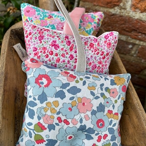 May include: Three floral-patterned sachets in a wooden bowl. The sachets feature pink, blue, and yellow flowers on a white background. A light gray ribbon is attached to the sachets. The tags read "Oxney" and "Olio Of".