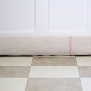 May include: A long, beige doorstop with red and white stripes. The doorstop is made of fabric and is lying on a chequered floor.