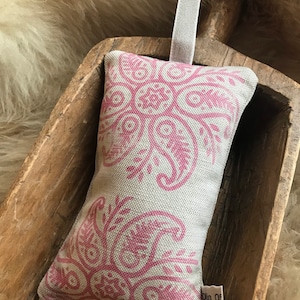Op de afbeelding: Een roze en witte stoffen zakje met een paisley-patroon. Het zakje heeft een lussen van linnen om op te hangen en een klein label met de tekst "Olio Of".