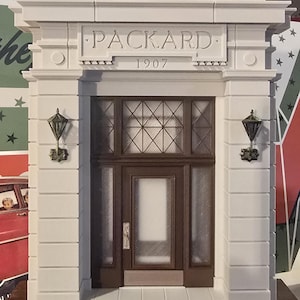 Può includere: Modello dettagliato di una facciata di edificio con il nome "PACKARD" e l'anno "1907" scolpiti sopra la porta. La facciata è bianca, con una porta e pannelli di vetro marroni. Due applique ornate fiancheggiano la porta.
