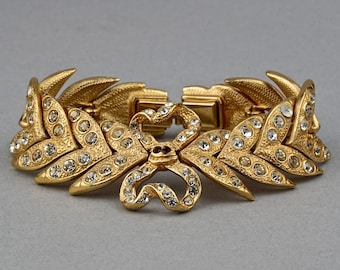 Puede incluir: Un brazalete dorado con un diseño en forma de lazo y brillantes transparentes. El brazalete está hecho de metal y tiene un cierre de bisagra.
