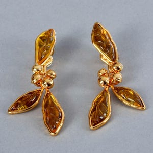 May include: A pair of gold-toned earrings featuring a leaf design with amber-colored accents. Each earring has three leaves and a cluster of small, round beads. The leaves have detailed veining, and the earrings are set against a gray background.