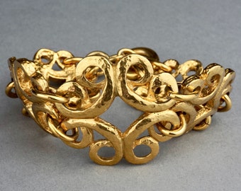 Puede incluir: Un brazalete dorado con un diseño ornamentado y texturado. El brazalete está hecho de lazos entrelazados que forman un patrón de remolino.