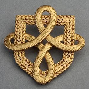 May include: A gold-toned metal brooch in the shape of a knot. The knot is made of braided metal and has a square shape.
