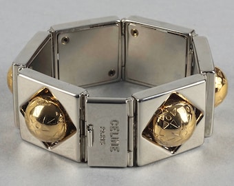 Puede incluir: Un brazalete de tono plateado y dorado con un diseño geométrico. El brazalete está hecho de eslabones entrelazados y presenta una pieza central redonda de tono dorado con un patrón geométrico. El brazalete está grabado con las palabras "CELINE PARIS".