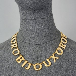 May include: A gold-tone necklace with large, chunky letters spelling out the word "BIJOUX" in French. The necklace is on a gray mannequin.