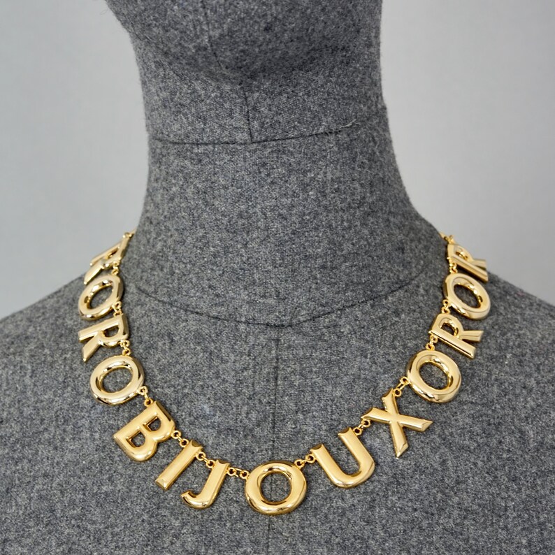 May include: A gold-tone necklace with large, chunky letters spelling out the word "BIJOUX" in French. The necklace is on a gray mannequin.