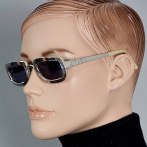 May include: Silver-toned rectangular sunglasses with dark lenses and a white plastic detail on the frame. The sunglasses have the letters "CD" on the side.