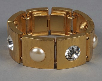 Puede incluir: Un brazalete dorado con cierre de bisagra. El brazalete está adornado con piedras redondas transparentes y blancas.