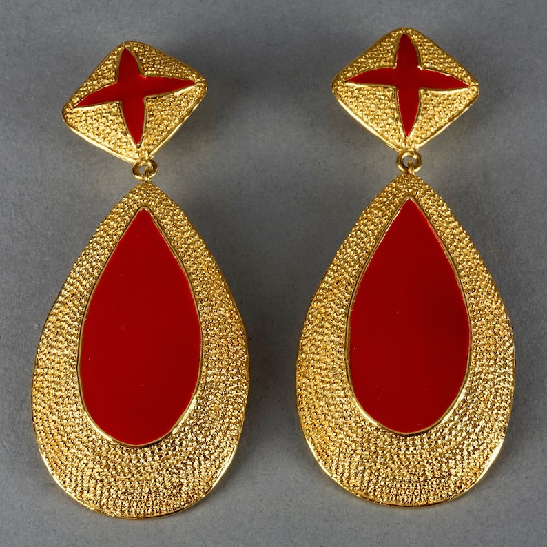 May include: A pair of gold tone dangle earrings with red enamel teardrop shapes and a cross design at the top.