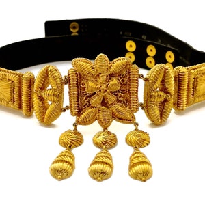 Pode incluir: Um cinto dourado com uma fivela decorativa e pingentes. O cinto é feito de metal tecido e tem um suporte de couro preto.