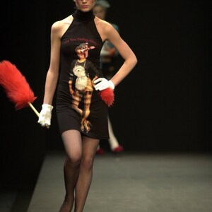 May include: A model on a runway wearing a black halter dress with a graphic print of a woman in a maid outfit. The dress has the text "Are You Feeling Dirty?" The model is holding a red feather duster and wearing white gloves.
