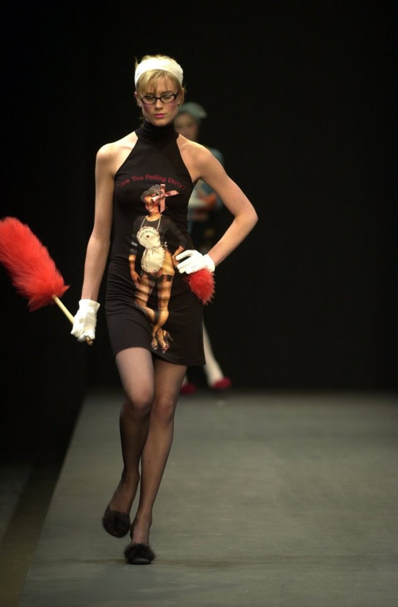 May include: A model on a runway wearing a black halter dress with a graphic print of a woman in a maid outfit. The dress has the text "Are You Feeling Dirty?" The model is holding a red feather duster and wearing white gloves.