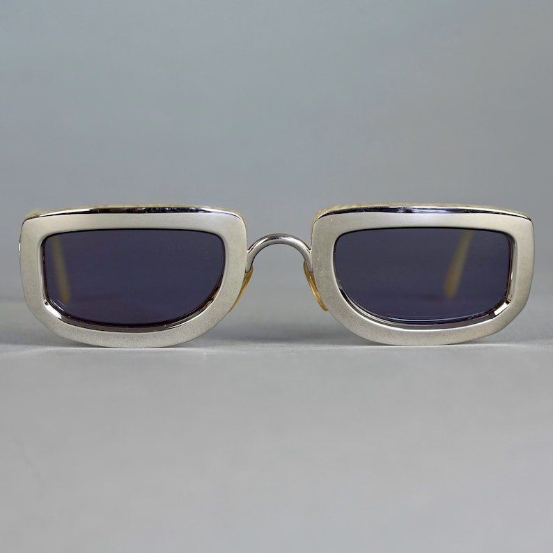 May include: Silver framed sunglasses with dark lenses. The frame has a subtle, textured pattern.