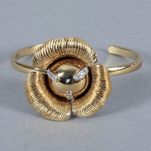 May include: A gold-toned cuff bracelet featuring a floral design. The center of the flower is a gold sphere, accented with small, sparkling gemstones. The petals are textured, and the bracelet has a simple, open design.