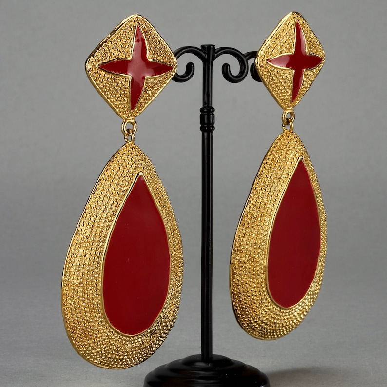 May include: A pair of gold tone dangle earrings with red enamel teardrop shapes. The earrings have a textured surface and a small red star on the top of each teardrop.