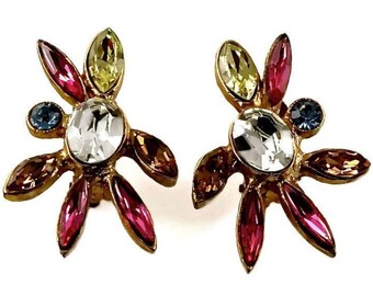 May include: A pair of gold tone earrings with a flower design. The earrings feature a large clear crystal in the center, surrounded by smaller crystals in shades of pink, blue, yellow, and brown.
