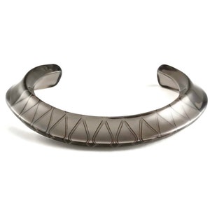 May include: A dark gray, semi-circular bangle bracelet with a geometric pattern etched into the surface.