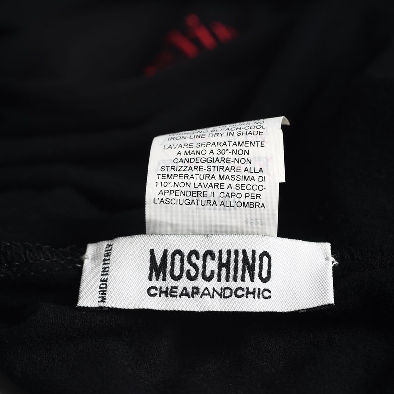 May include: Close-up of a black garment with two white tags. The top tag has washing instructions in Italian. The bottom tag reads "MOSCHINO CHEAPANDCHIC" with "MADE IN ITALY" printed on the left side.