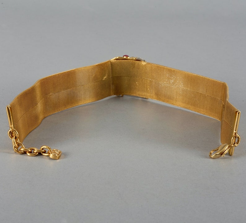 May include: A gold mesh choker necklace with a red and clear stone embellishment. The necklace has a clasp closure.