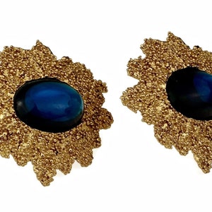 May include: A pair of gold earrings with a blue gemstone in the center of each earring. The earrings are shaped like a flower with a textured surface.